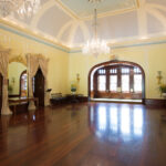 Image: A large, open room with a wooden floor, yellow painted walls, an ornate ceiling, and portraits hanging on the walls