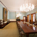 Image: A large room containing a long table with chairs. Two large portraits and a mirror hang on the walls