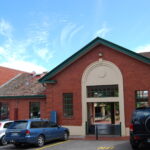 Image: The front of a single-storey brick building with a white arch entranceway. Several modern auto-mobiles are parked in front of the building