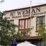 Image: The upper floor of a rectangular building with two windows and a set of double-doors. The words ‘Hy. Weman. Sail Maker, Ship Chandler’ are painted on the building front