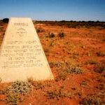 Image: A pyramidal-shaped concrete plinth stands in a vast, open desert area. A notation inscribed on the plinth warns of radioactive contamination