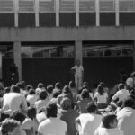 Image: A man in white stands with a microphone in front of a group of seated students