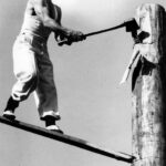 Image: man holding large axe standing on wooden plank embedded in large wooden post