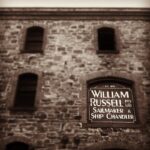 Image: Close-up photograph of part of an historic, multi-storey stone building. The words ‘Est. 1870, William Russell Pty. Ltd., Sailmaker and Ship Chandler’ are painted on the shutter of one of the building’s second-floor windows