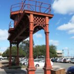 Image: A nineteenth-century iron gantry crowned by a wooden observation tower forms an archway over a modern carpark. An historic marker on a stone plinth is positioned next to the gantry