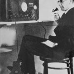 Image: A middle-aged clean-shaven Caucasian man wearing a suit and wire-rimmed spectacles sits in a chair next to a desk. On the desk is a large pedal-powered radio, the handset of which is held by the man