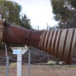 Image: The rusted exhaust section of a rocket mounted on a pedestal near a cluster of gum trees