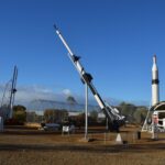 Image: Several different rockets are exhibited on pedestals on a flat, scrubby expanse of ground