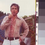 Image: Man in pink paisley shirt and white trousers speaks into a microphone