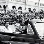 Image: Black and white photo of people in car surrounded by a crowd