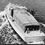 Image: A boat with a group of children sitting in the stern travels along a waterway. Two policemen are visible standing at midships on the starboard side