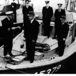 Image: Six men stand on the bow of a boat. One man in a naval uniform passes a paper document to another man in a civilian suit