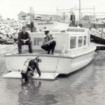 Image: A scuba diver exits the water at the stern of a small motor boat. Two police officers aboard the boat look on