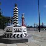 Image: A large plinth painted with an alternating black and white pattern, and emblazoned with the words ‘Keep Left’ and directional arrows at its base. It is displayed on a street corner. A red lighthouse tower is visible in the background