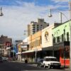 Image: a photograph of modern city street with two storey terraces painted in a range of bright colours in the foreground and skyscrapers in the background. Most of the shops are currently being used as restaurants, bars and nightclubs.