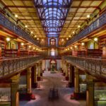 Image: The interior of a large, three-storey Victorian-era library. A large skylight in the roof illuminates the building’s three floors of bookshelves and museum exhibits