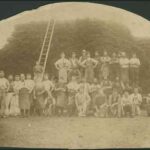 Image: A large group of men in working clothes, some wearing aprons, pose for a photograph in front of a ladder leaning on a large stack of wattle bark. To the right of the photograph, one man kneels holding a small dog.