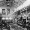 Image: a large open hall with a second storey gallery with flags hung from the railing. Rows of chairs and glass cabinets displaying various items fill the ground floor.