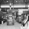 Image: a number of displays in glass cabinets fill a large hall. In the centre of the photograph a display of varnishes is topped by a pyramid. To the left a man stands in a uniform of a dark jacket and white pants and hat.