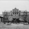 Image: A three storey building with a large external staircase leading down multiple terraces to a lawned area planted with trees and flowers. At various places on the staircase men stand in uniforms of dark coats and white pants.