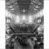 Image: a pipe organ takes pride of place at the far end of a large hall with a hammer beam roof. In the foreground men and women in black sit on rows of chairs surrounded by glass display cabinets.