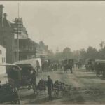 Image: A boy holding two hand-carts stands to the side of a busy street surrounded by horse-drawn carts. A tram line runs down the middle of the street, stone and brick buildings, one with a large balcony, line one side while the other fronts parklands.