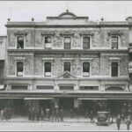 Image: a three storey stone terrace building with a verandah and triangular pediments over the rectangular windows.