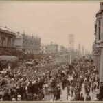 Image: a large crowd of people watch a contingent of soldiers in late 19th century military uniforms march down a wide city street led by four men on horseback