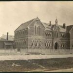 Image: a large brick building with arched windows and doors. Lighter coloured bricks are arranged in decorative diamond patterns between the first and second storeys and in an alternating pattern with the darker bricks on the arches above the windows.