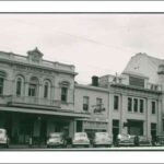 Image: a row of 1950s era cars are parked on a street outside of a row of two and three storey commercial buildings including a hotel with a verandah and balcony and a bank with a domed roof and flag pole.