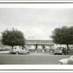 Image: a number of 1940s and 1950s era cars are parked in rows outside a single storey building with a sign reading "used car division"