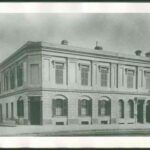 Image: a two storey stone building with corner entrance, arched windows with shutters on the first floor and rectangular windows above