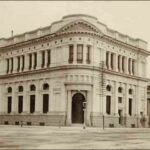 Image: a two storey stone building with a curved corner.The entrance on this corner is flanked by columns. The ground floor windows are arched while the upper floor windows are rectangular and arranged in pairs except for the corner which has a group of 3