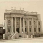 Image: a three storey stone building with twin curved staircases leading up to its main entrance which is flanked with two storey high columns. The roof is decorated with a balustraded parapet with decorative urns.