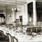 Image: a large dining room with multiple tables set with white cloths, candelabras and floral centrepieces.