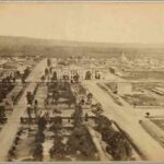 Image: an aerial photograph of a city showing a range of large public and smaller residential buildings in the distance and a large public garden and empty plots in the foreground