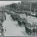 Image: men wearing Second World War era military uniforms and carrying rifles parade down a wide city street whilst crowds watch on from either side.