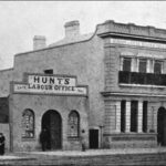 Image: a simple two storey building with an arched door to one side, a balconet and a small parapet stands next to a single storey building with a central arched door flanked by arched windows and a parapet sign reading Hunt's Labour Office