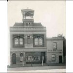 Image: A narrow two storey building with a Queen Anne style turret rising from the centre of the building's frontage. Recessed arched windows on the second floor allow for small balconies with decorative fretwork.