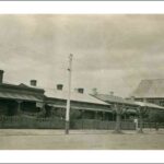 Image: a street-scape featuring a row of single storey cottages with tin roofs and verandahs behind picket fences. At the far right of the image a small stone church can be seen. The street is also lined with heavily pruned trees and telegraph poles.