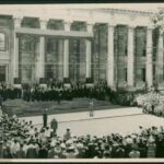 Image: a man stands in front of a wide staircase leading up to a grand building with columns, photographing the men who are arranged upon it. A large crowd surrounds him.