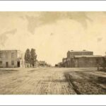 Image: The intersection of two wide dirt roads. The nearest corners are fenced with grass and low lying shrubs. The far left corner supports a two storey hotel and the far right a collection of stone buildings, one with a sign reading "Timber Yard"