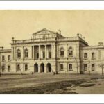 Image: a large stone public building with a three arched entrance above which is a balcony with columns. A smaller third storey with a gable roof rises from the centre of the building. Two storey wings extend to either side.