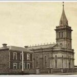 Image: A stone church with a square central spire. To the left is a simple, rectangular two storey building with a central door flanked by windows on each side on the ground floor and three windows in line with the openings below on the second floor.