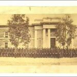 Image: a large group of men in 1860s police uniforms stand in front of a two storey stone building with columns flanking its entrance.