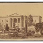 Image: three young girls walk through a garden in front of a two storey stone building with a portico featuring a triangular pediment supported by four columns