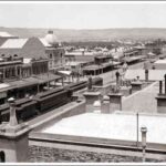 Image: a train is stopped in the centre of a city street which is lined with two and three storey buildings most of which have verandahs and one which features a large domed roof.