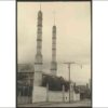 Image: two minarets front a mosque partially obscured behind a high wall and trees