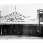 Image: a single storey shop with a verandah and triangular parapet with a painted sign reading "Lenroc Limited Central Service Station for Harley-Davidson, Triumph & Raleigh motor cycles & side cars". A flag flies from the peak of the parapet.