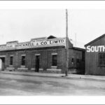 Image: a single storey brick building with a low pitched roof behind a parapet sign reading "Balfour, Bricknell & Co. Ltd. Cake Specialists". To the right is a corrugated iron building with part of a sign visible reading "South Aust"